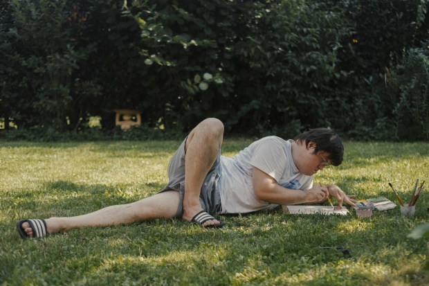 Zhenya Holubentsev liegt auf einer Wiese und malt. Ein Bein hat er angewinkelt, das zweite flach ausgestreckt.