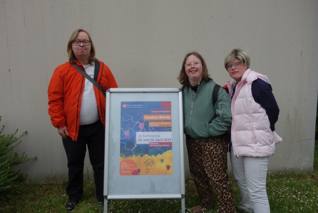Ein Mann und zwei Frauen mit Down-Syndrom stehen recht und links von einem Aussteller mit einem Plakat mit dem Schrifttzug Gelebte Würde.